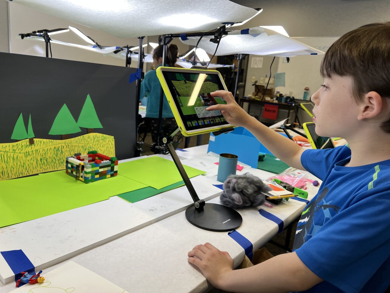 Color photograph of a child in a blue shirt using an iPad to film a lego building against a hand drawn set with trees and grass.