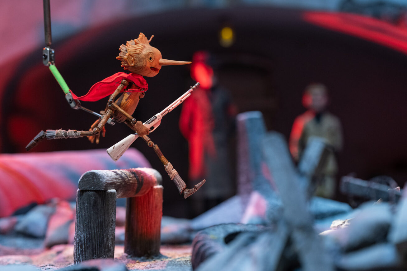 Close-up photograph of the Pinocchio puppet wearing a cape and holding a rifle while leaping over a barricade.