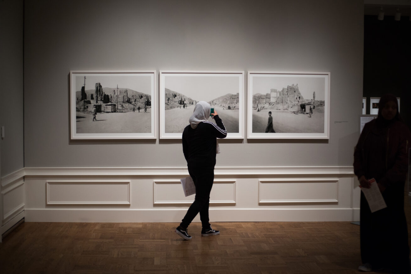 Person wearing a white headscarf looking at three black and white photographs in a dimly lit gallery