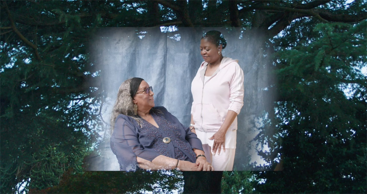 Two older black women looking at each other, one sitting and one standing. Behind them is the image of tree branches.