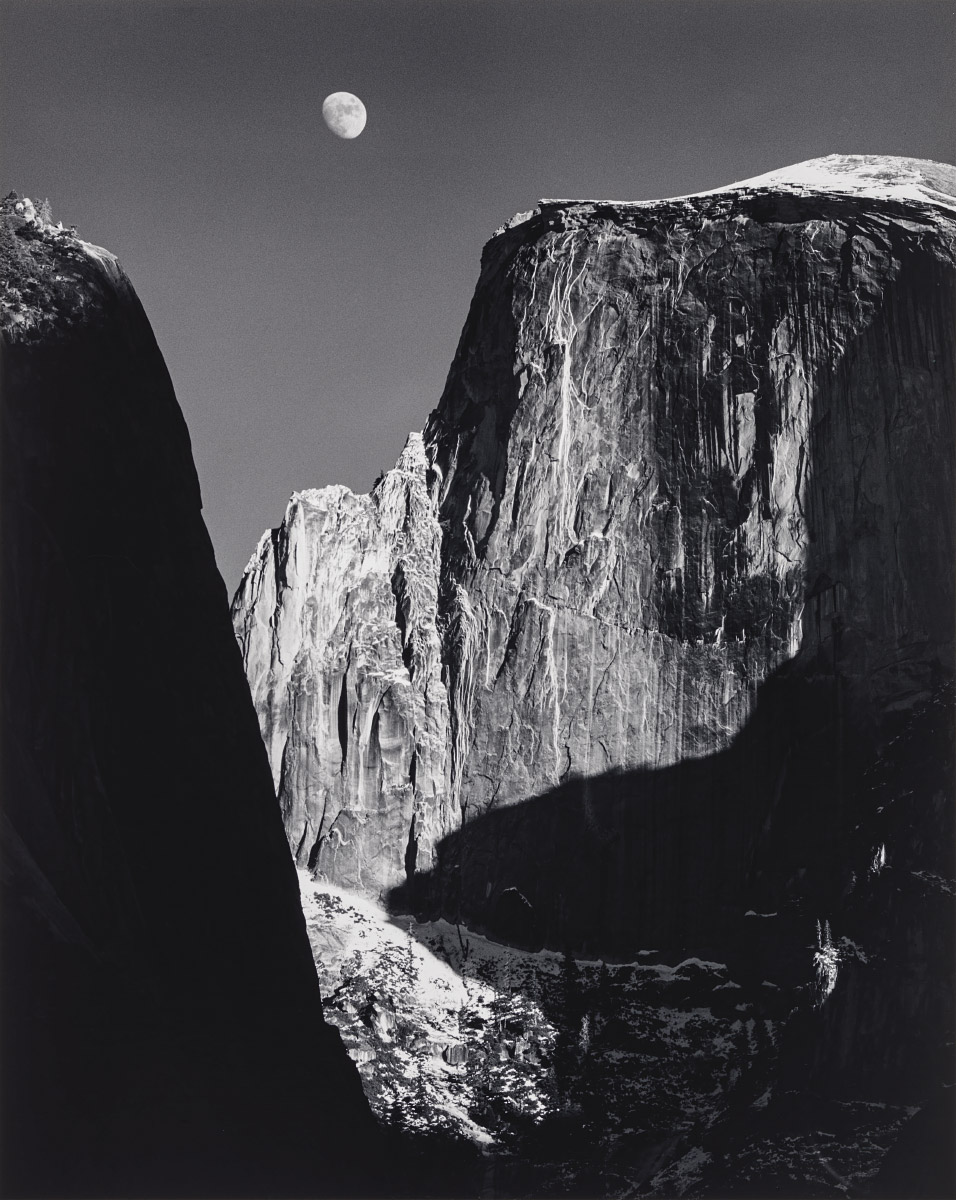A black and white photograph through a canyon at Yosemite National Park to a view of Half Dome and the moon in the middle