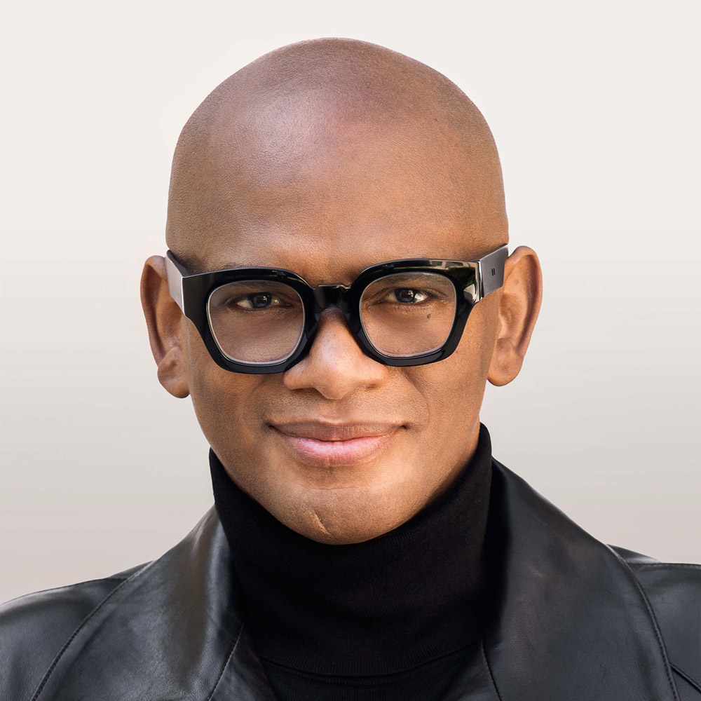 Portrait of a Black man who is bald, wearing heavy black eyeglasses, a black turtleneck, and a black leather jacket