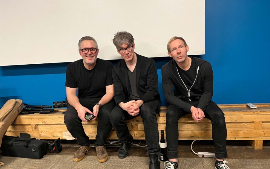 Three men wearing all black sitting on a wooden bench against a blue wall with a whiteboard on it