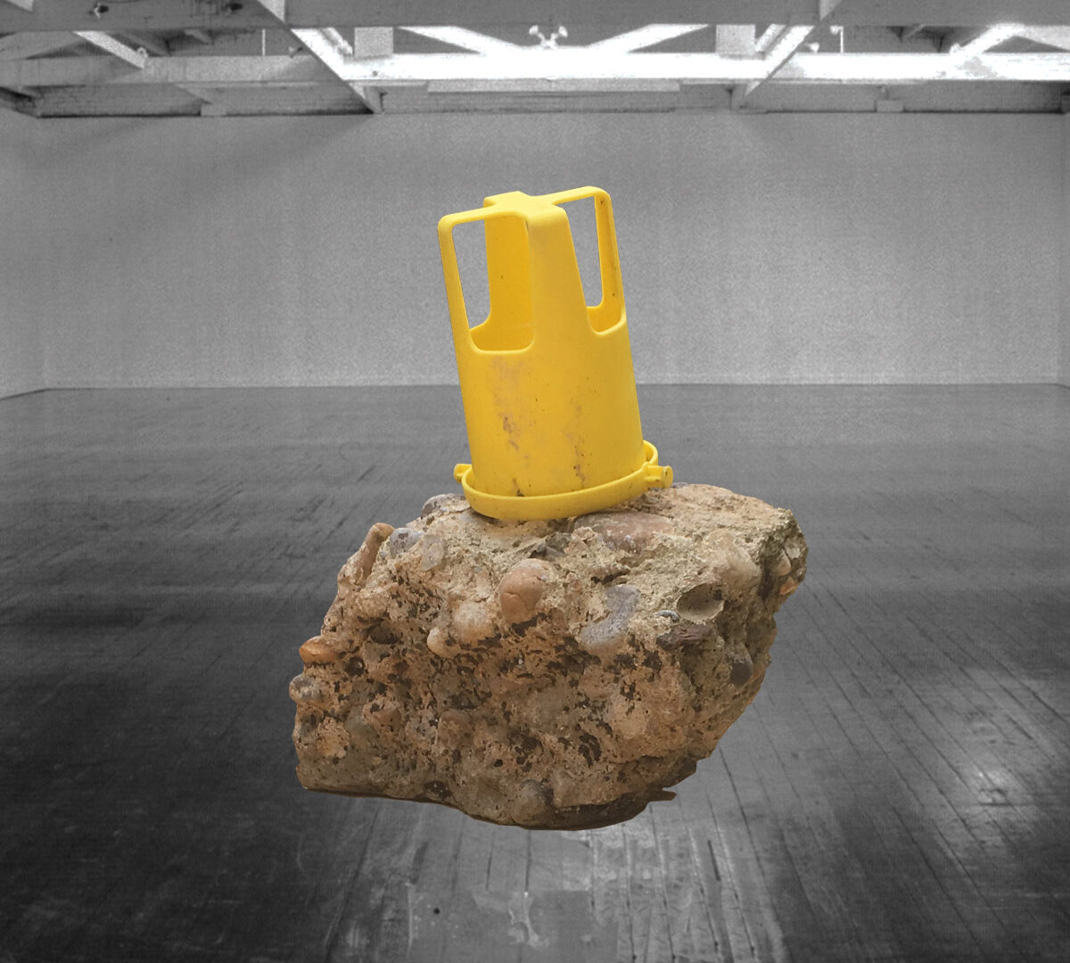 Sculpture of cement and rock with a yellow plastic item on top, hanging in a white-walled gallery with a black floor