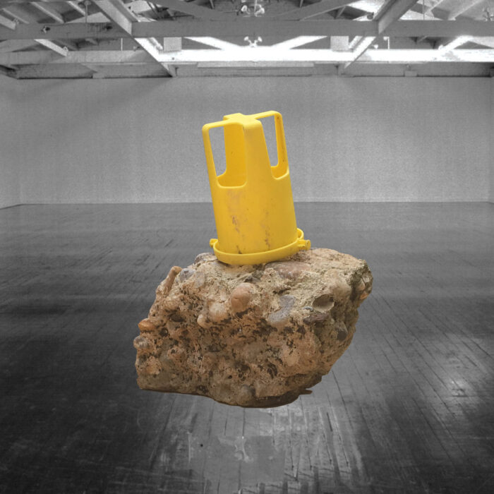 Sculpture of cement and rock with a yellow plastic item on top, hanging in a white-walled gallery with a black floor