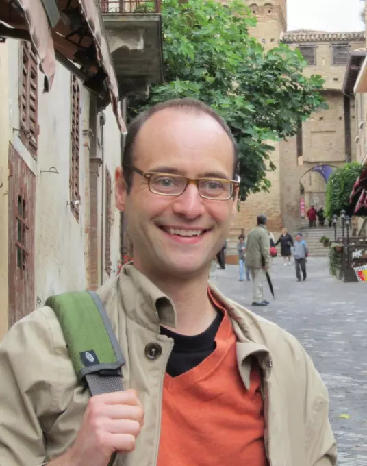 A smiling man with eyeglasses wearing a khaki jacket over orange and black shirts. Standing on a cobblestoned street.