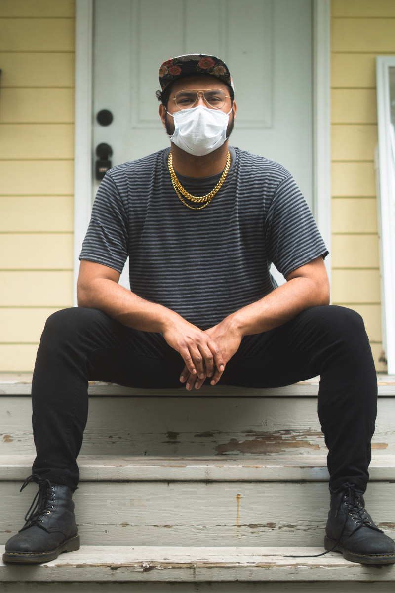 A person sitting on the steps in front of a yellow house with a white door. They are wearing a mask, a grey shirt, black pants, and black combat boots and have their legs spread out in front of them.