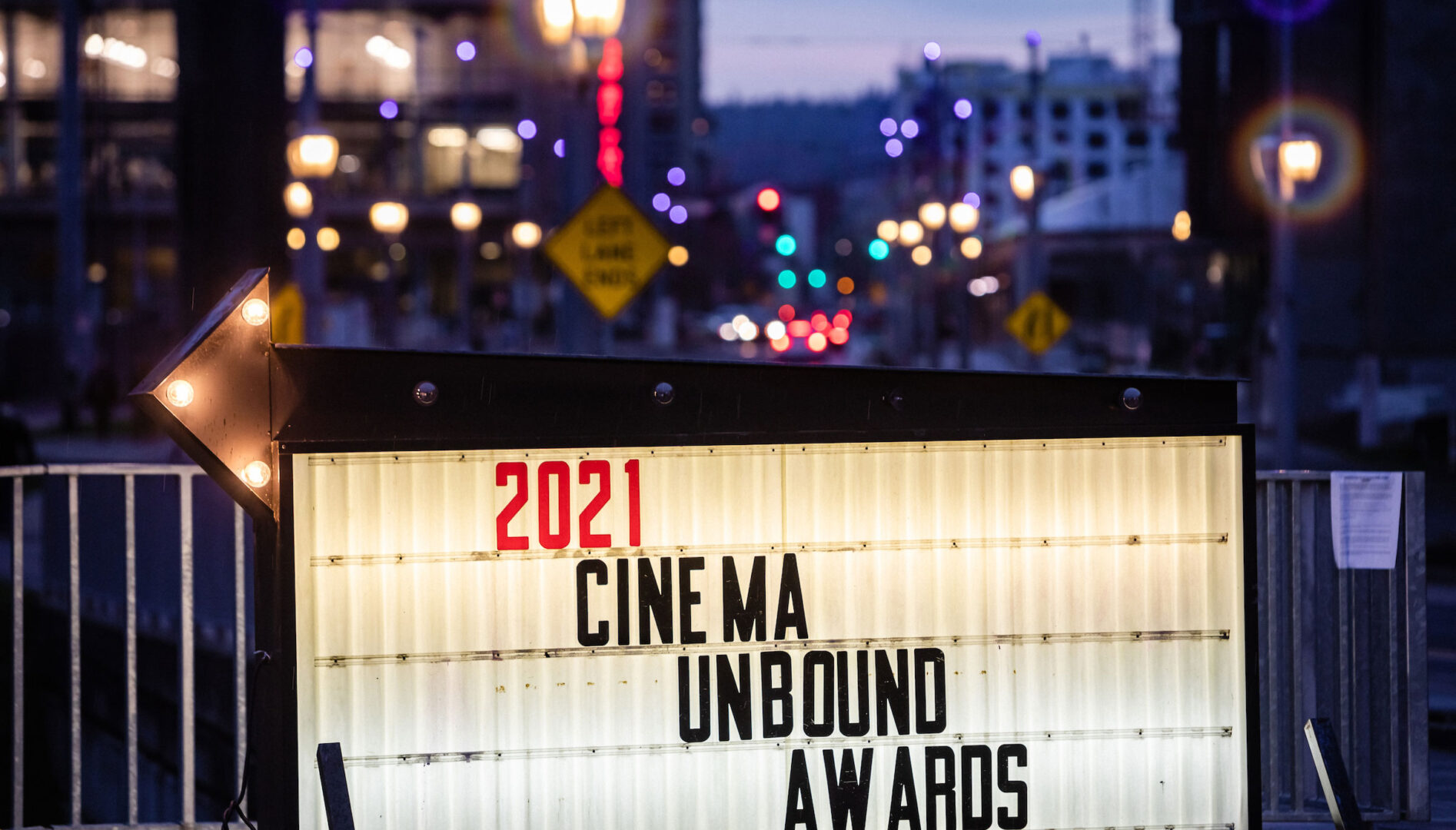 Lit up sign that says "2021 Cinema Unbound Awards" at dusk with cityscape and lights behind it