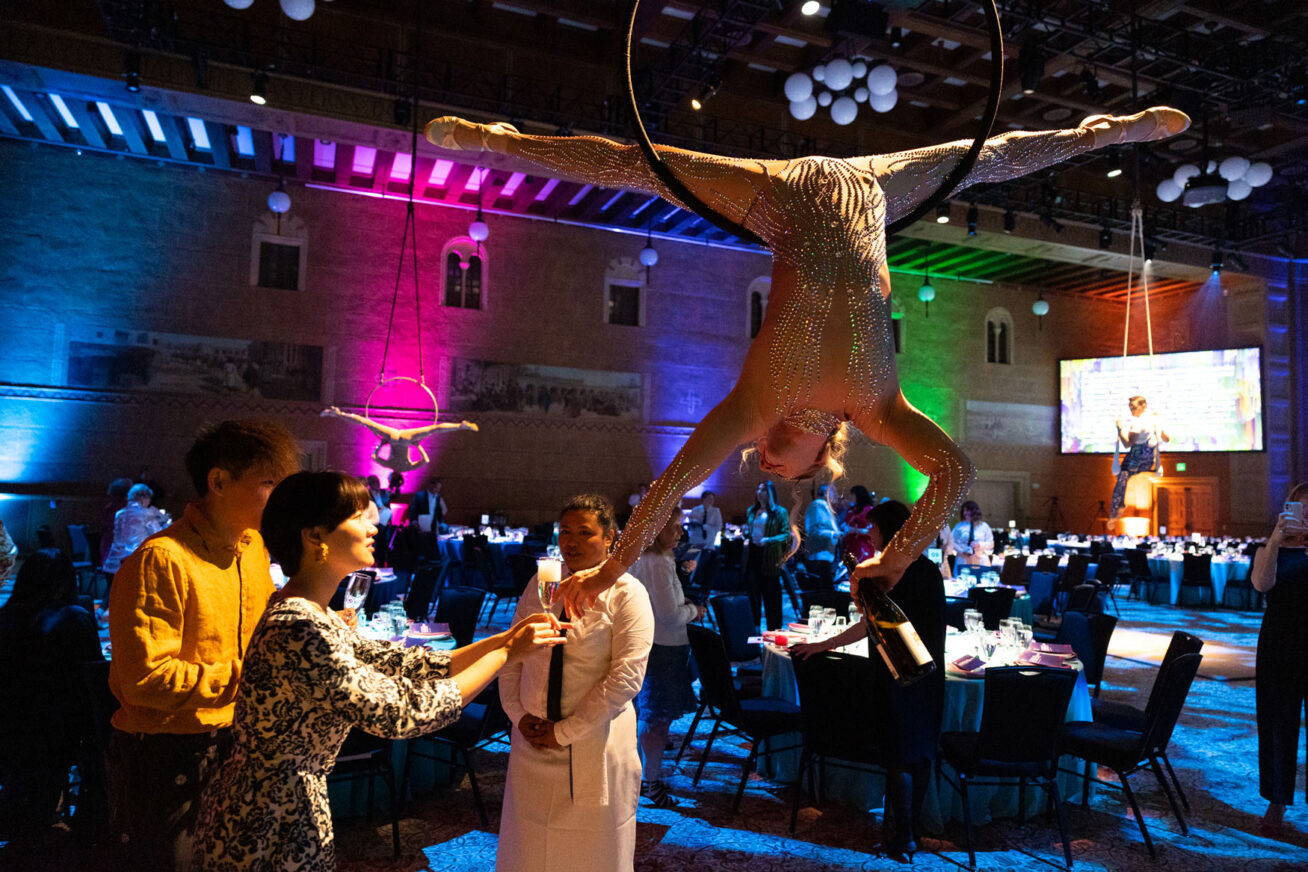 Aerial acrobat hanging upside down from a large ring and pouring champagne into a partygoer's glass