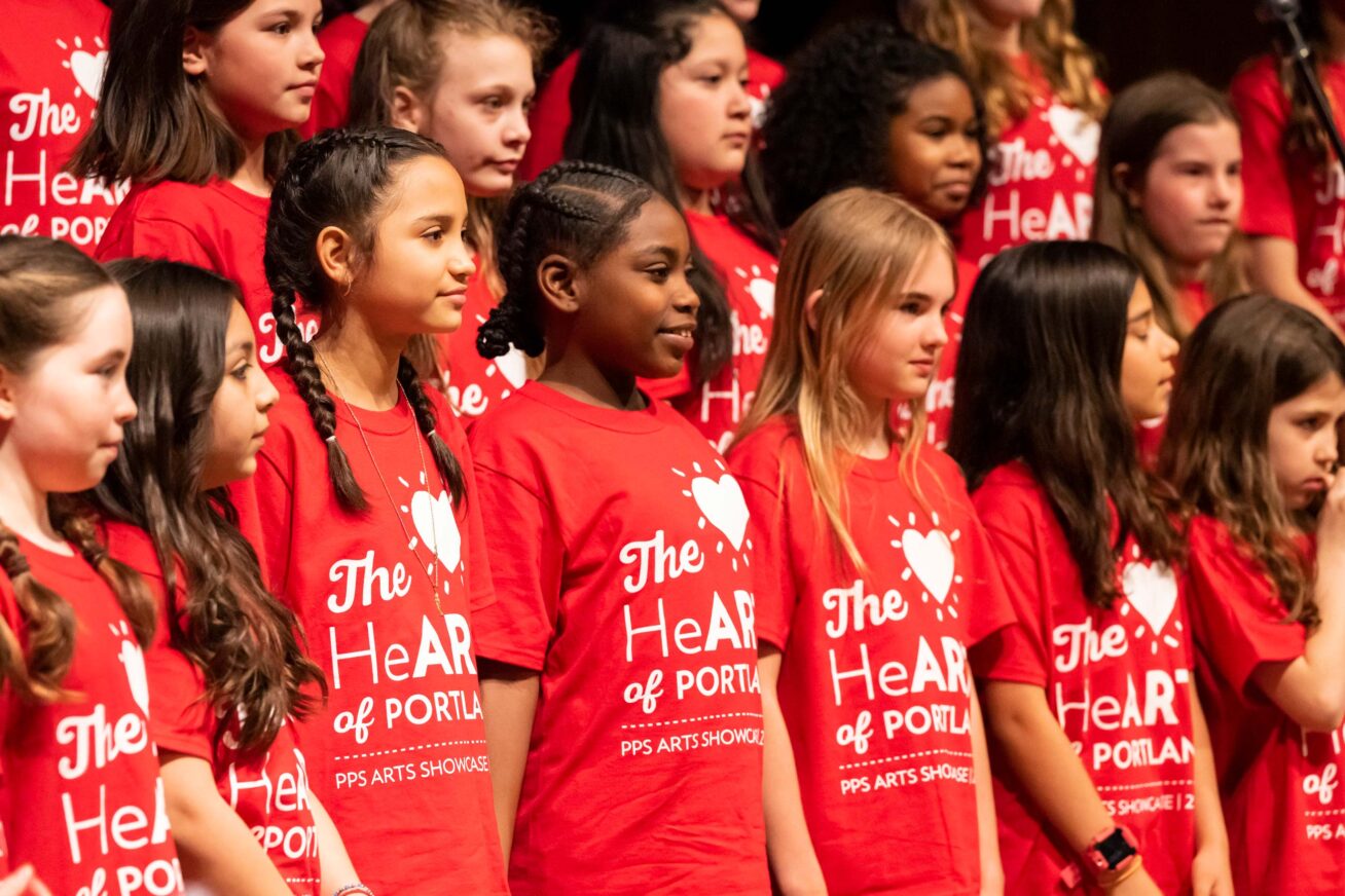 Group of Portland Public School students on stage in red HeART of Portland t-shirts smiling out towards a crowd