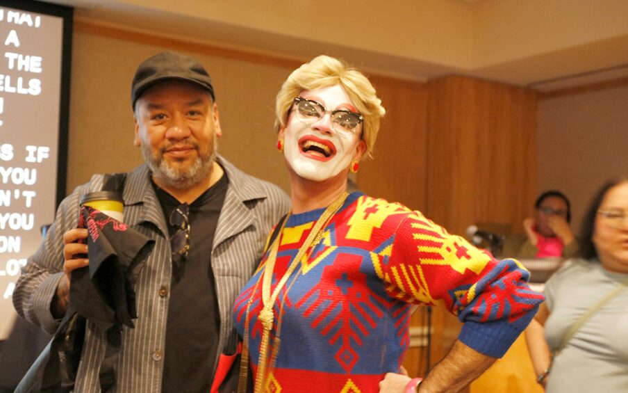 Man in a black hat with a grey beard, black t-shirt and grey and white striped coat standing next to a drag queen with a blond wig, white face paint, and a blue, red, and yellow sweater.