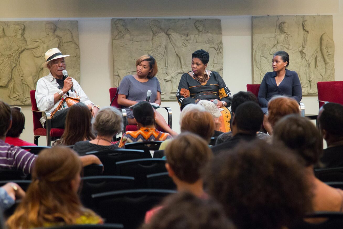 Group of four Black speakers at the Race in America After Ferguson discussion at the Museum. One man and three women, sitting on stage with an audience in front of them. The man, wearing a white hat, is holding a microphone and speaking.