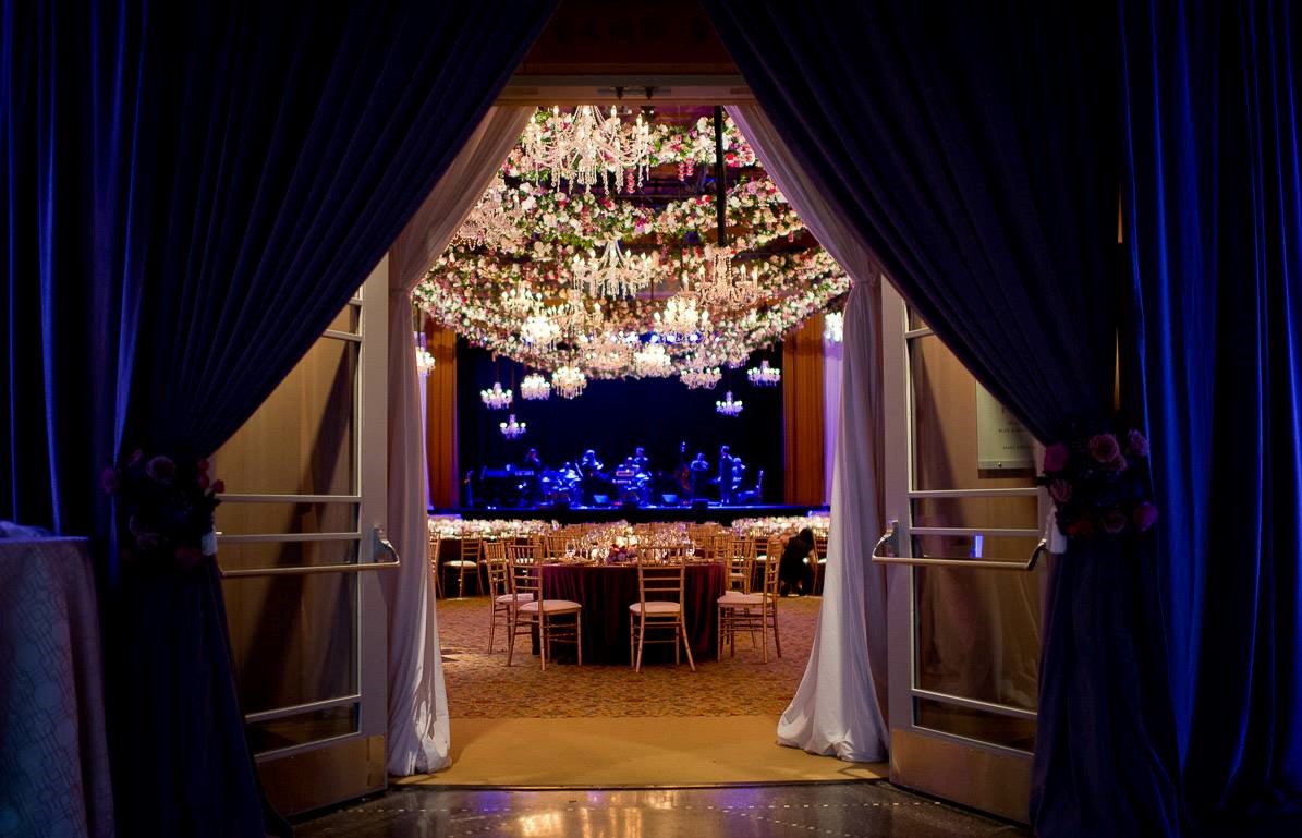 Color photo looking through parted blue velvet curtains into a ballroom with chandeliers and flowers hanging from the ceiling. Two doors are beyond the curtains, framing a view of set tables with chairs and a dimly lit stage in the background.