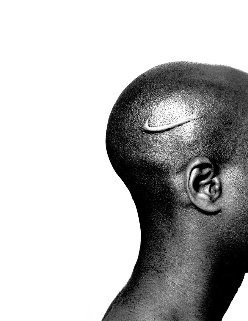 Black and white photo of a Black person's shaved head, ear, and neck with a branded Nike logo on their scalp