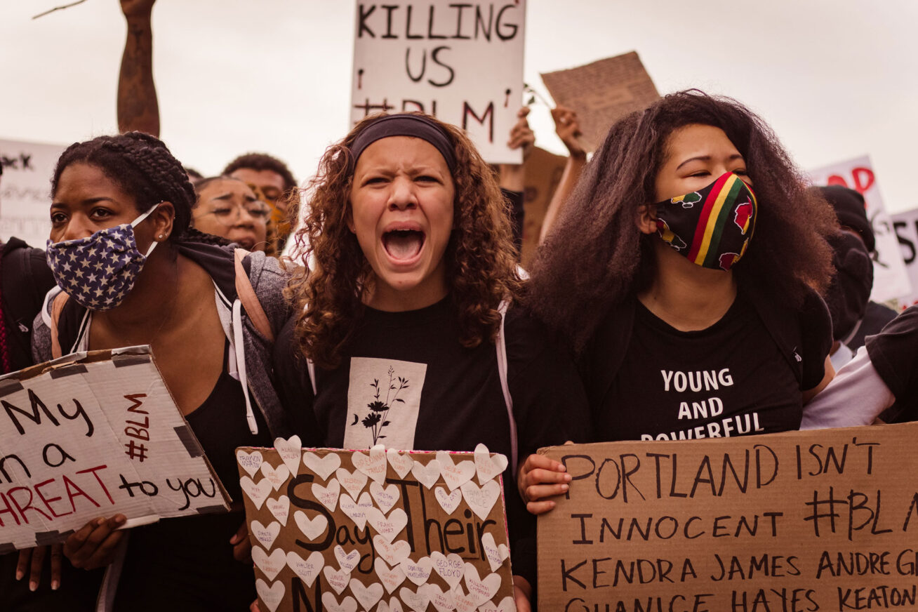 A group of protesters with three young women at the front, holding signs. The one in the middle looks like she's screaming.