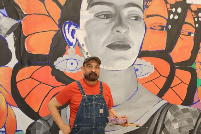 A man in overalls, a red tee shirt, a black baseball hat, and eyeglasses standing in front of a mural of Frida Kahlo with butterfly wings behind her.