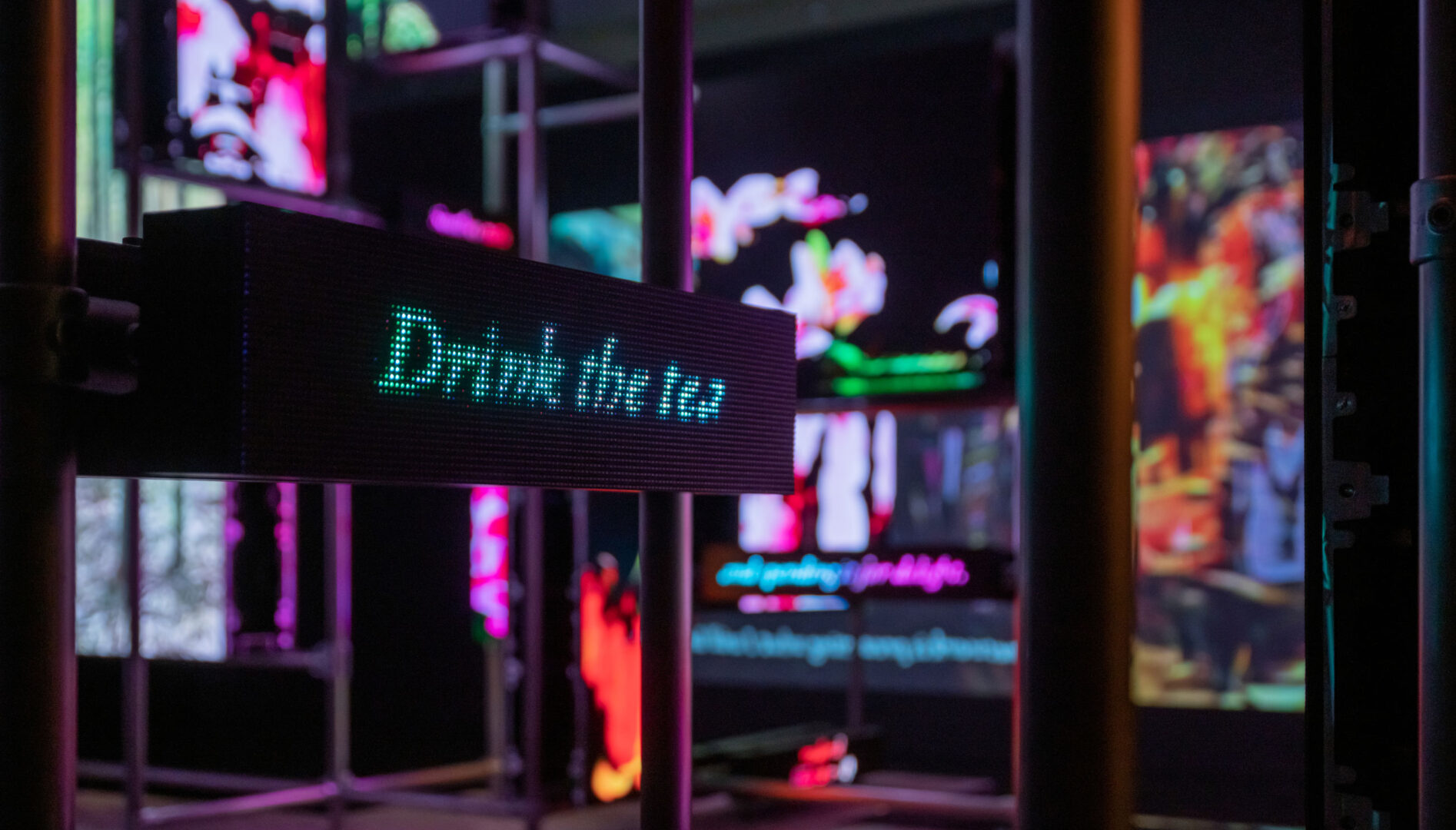 A dark gallery with various screens showing AI flowers and a pixelated screen that says "Drink the tea" in the foreground