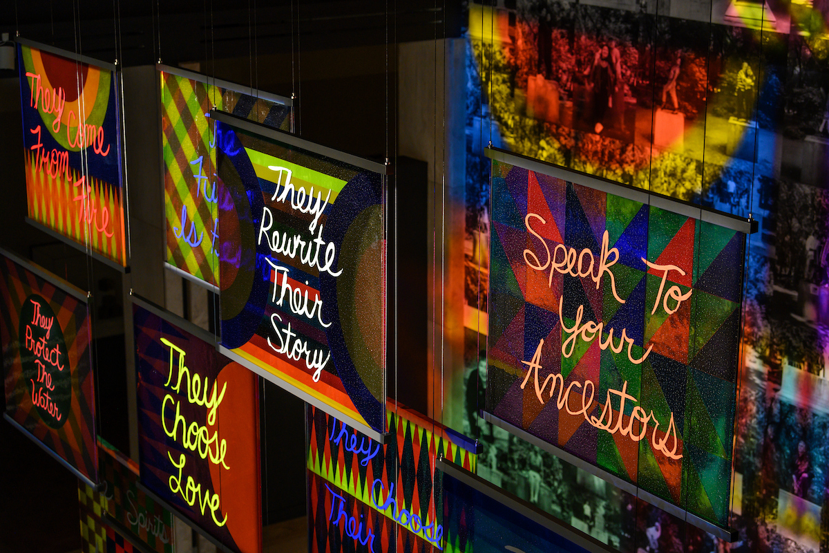 Suspended glass panels with hand painted text such as "Speak to your Ancestors", "They Rewrite Their Story", and "They Choose Love"