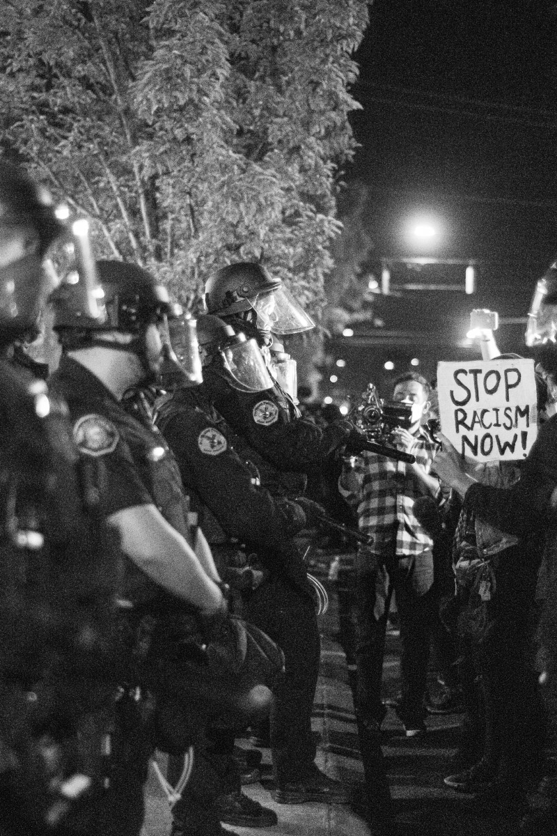 Black and white photo of a line of police wearing face shields to the left. At the end of the line is a person holding a video camera and a sign that says "Stop Racism Now."