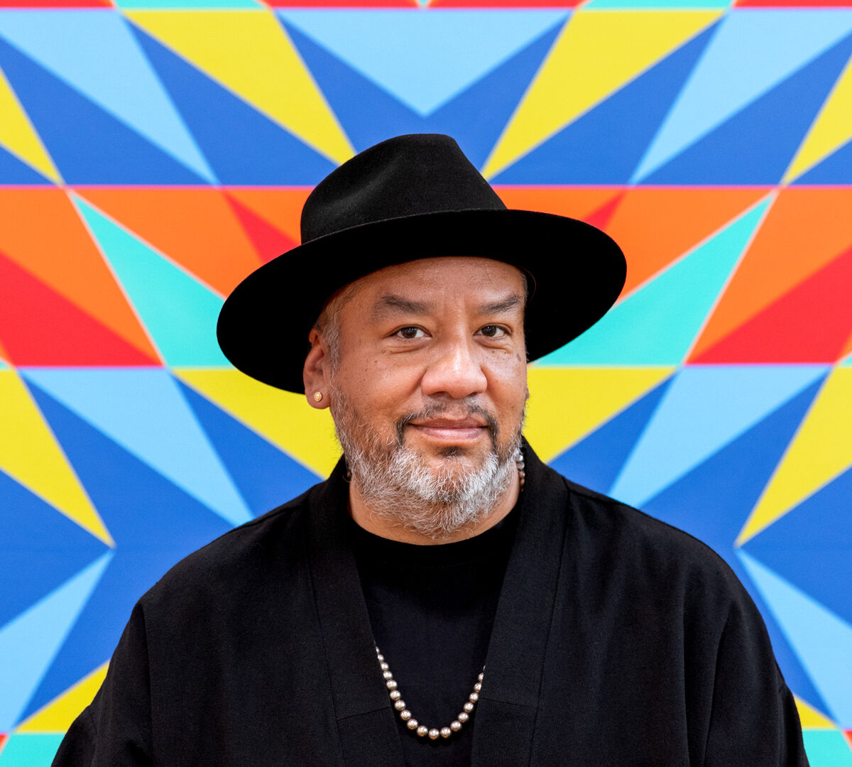 Portrait of artist Jeffrey Gibson wearing a black hat, a black shirt and jacket and a silver necklace. Standing against a colorful background.