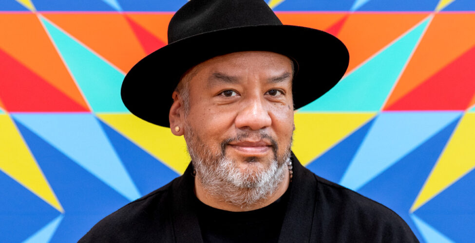 Portrait of artist Jeffrey Gibson wearing a black hat, a black shirt and jacket and a silver necklace. Standing against a colorful background.