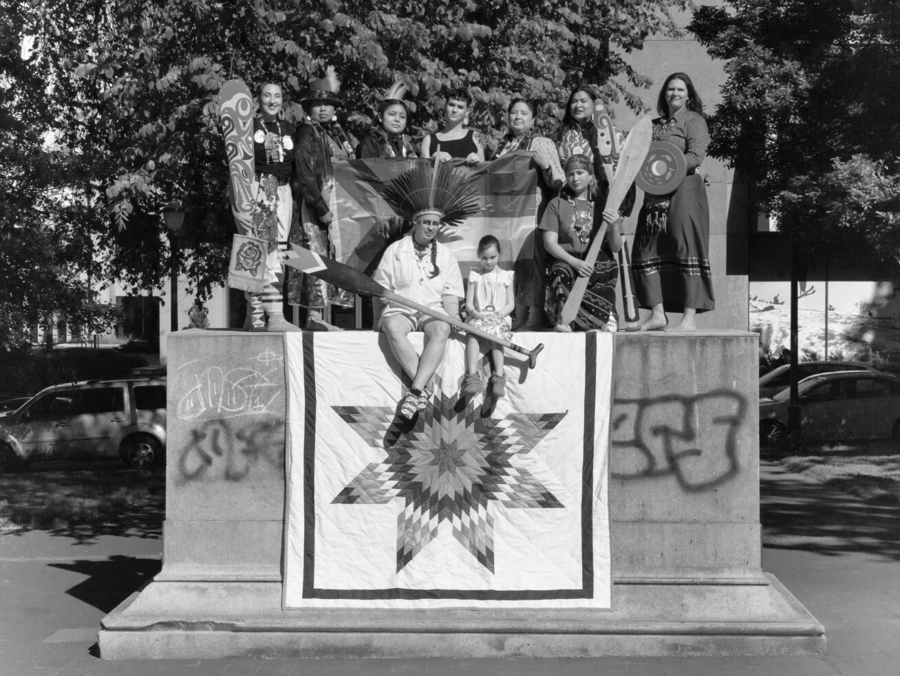 Black and white portrait of a group of Native people on a pedestal with the back row people standing holding a flag and a star quilt in front of them