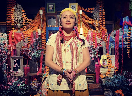 A woman with long hair and a yellow headscarf standing in front of a Dia de Muertos altar.