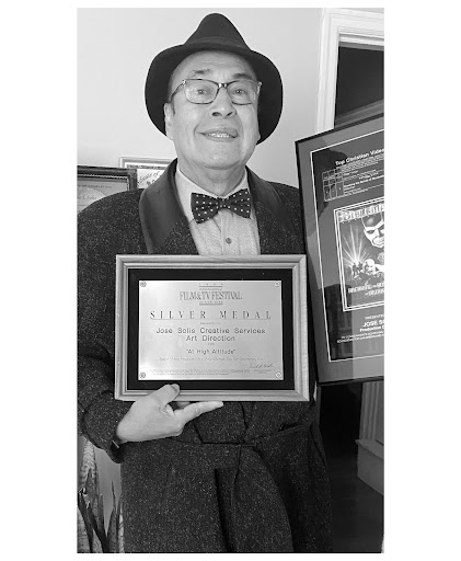Black and white photo of a smiling man in a fedora, a bowtie, a blazer, and eyeglasses holding up an award in front of him.