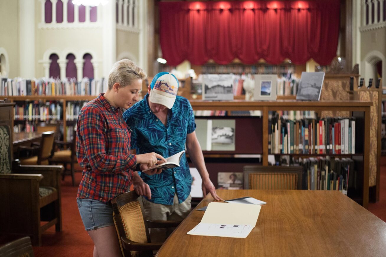 Two people looking at books at a wooden table. One with short blonde hair wearing a red plaid button down shirt and the other in a trucker hat and a blue Hawaiian button down shirt. Books shelves, old wooden chairs and a red curtain are behind them in the Crumpacker Library.