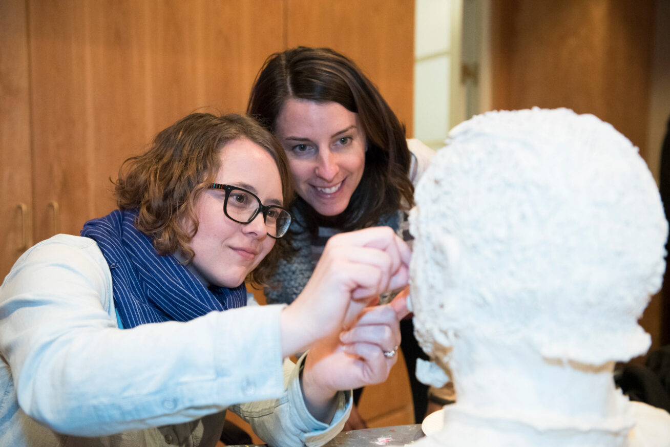 Two women, one with curly red hair and eyeglasses, one with long dark brown hair, working on a white sculpted head artwork