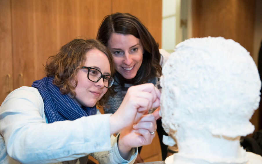 Two women, one with curly red hair and eyeglasses, one with long dark brown hair, working on a white sculpted head artwork