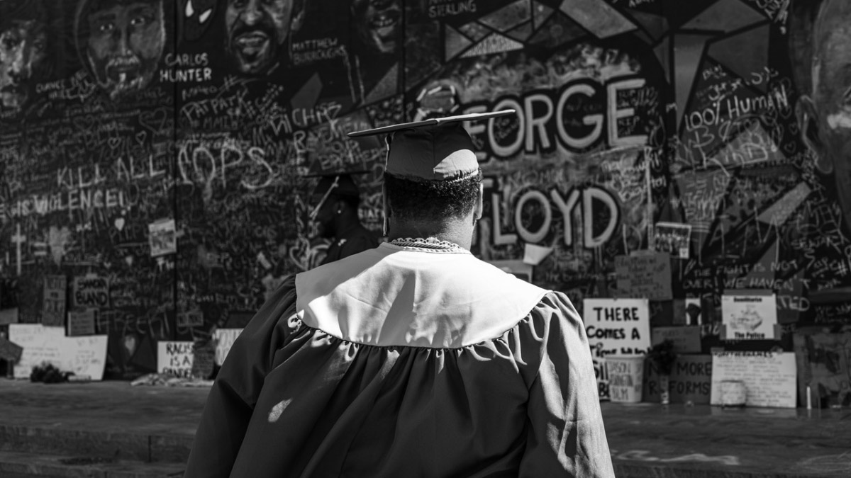 Black and white photo of person in a graduation gown and cap facing a wall of graffiti.