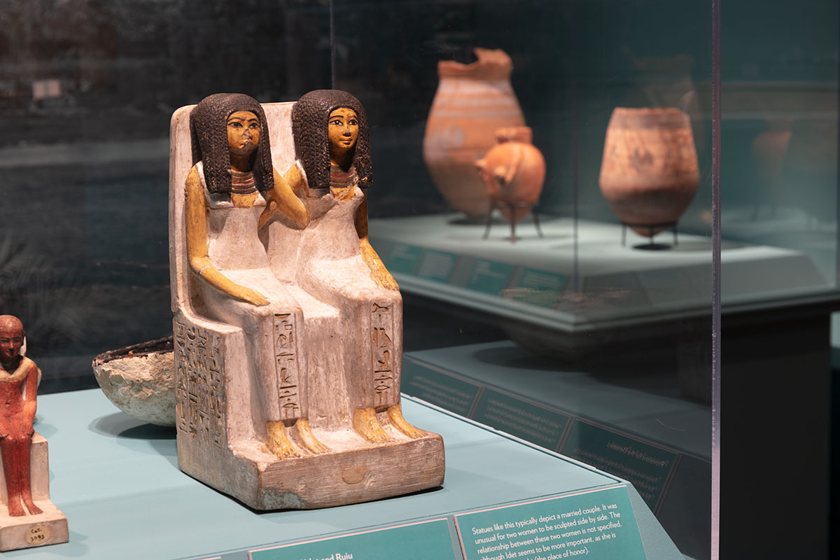 Statue of two Egyptian women with long brown hair sitting next to each other with their arms behind each other's backs