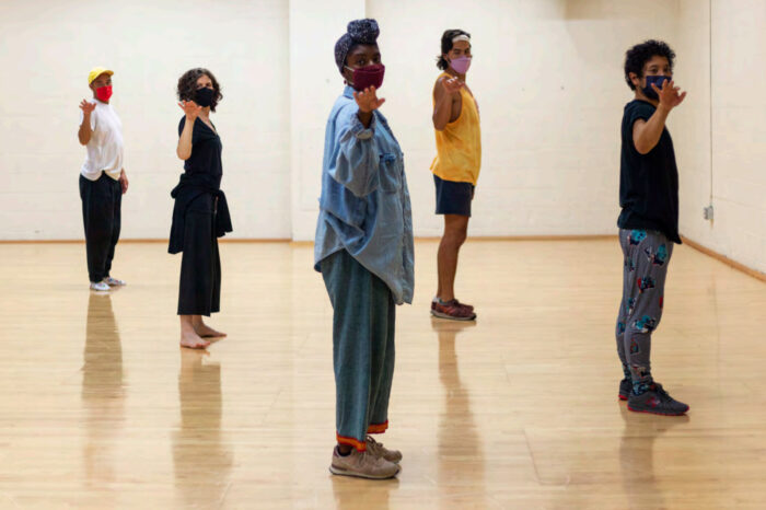 Five dancers in a studio with a light wood flood and white walls, wearing face masks, standing sideways with one arm reaching out towards the front.