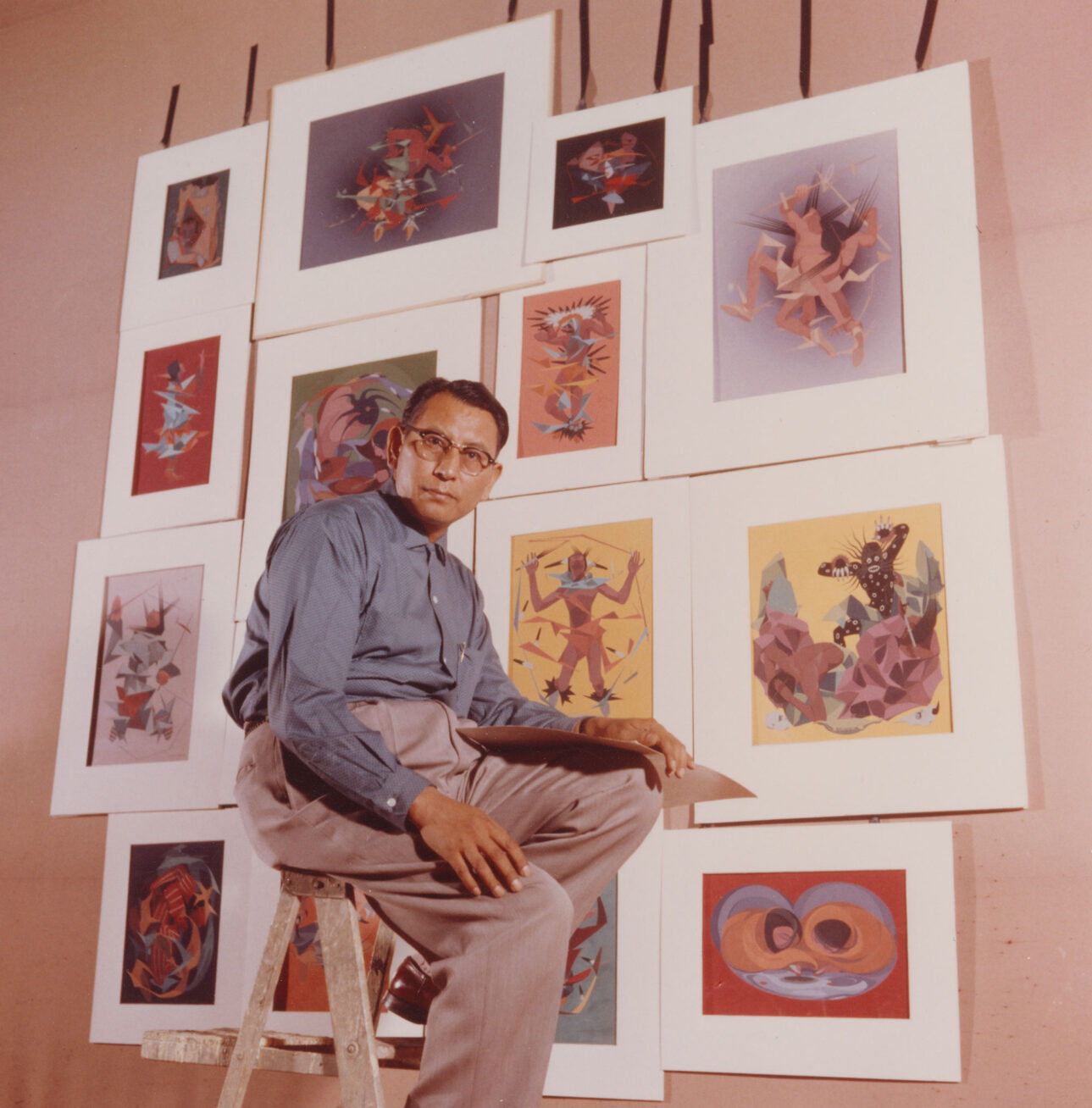 Artist Oscar Howe sitting on a ladder in front of a wall of his paintings.