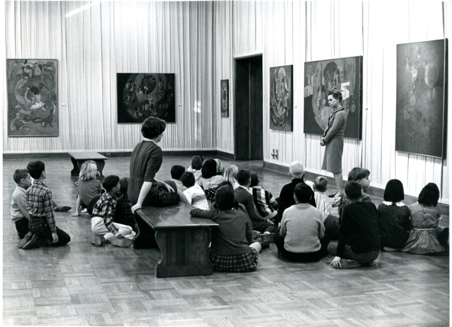 Historic, black and white photo of children sitting on the floor in an art gallery.
