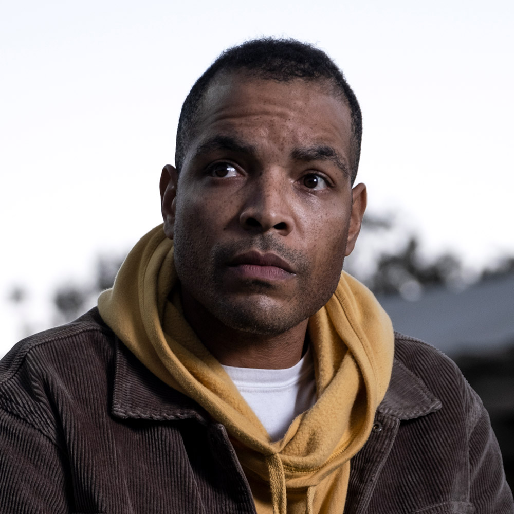 Portrait of a Black man looking to the side wearing a yellow hoodie, a brown corduroy jacker and a white t-shirt