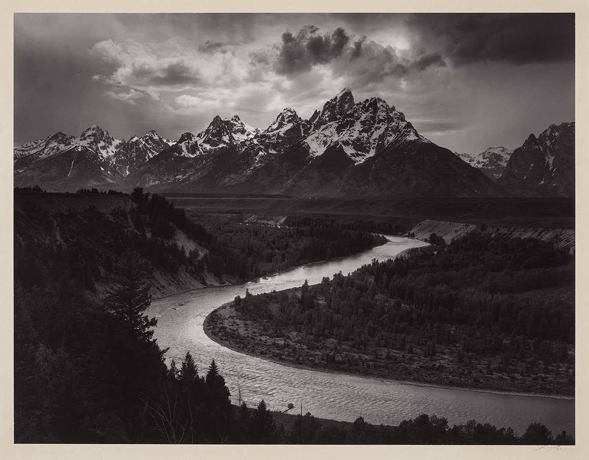 A black and white photo of a river bend with trees in the foreground and mountains in the distance