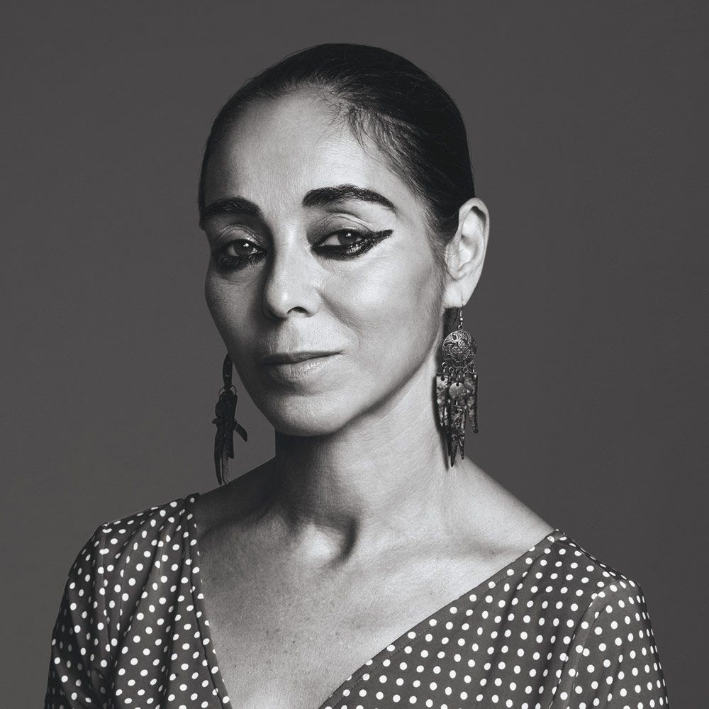 Black and white portrait of a woman with her hair pulled back wearing a v-neck, polka-dotted blouse, long metallic earrings, and heavy black eyeliner