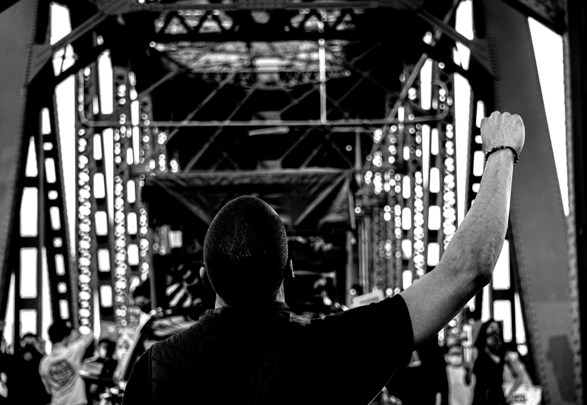 Black and white photo of a man's back. He is raising his right fist in the air and a crowd of people stand on a bridge in front of him.