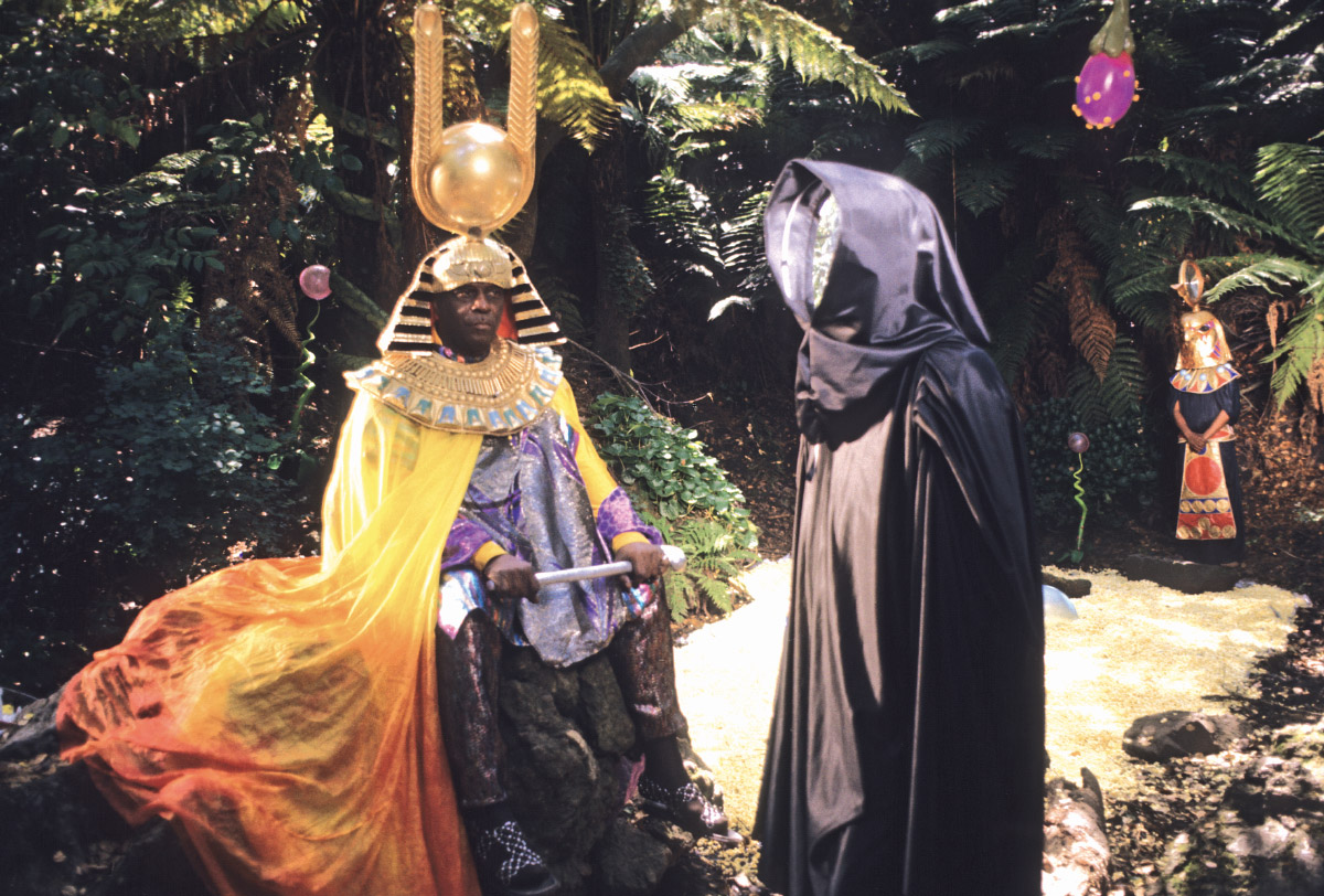Film still of Sun Ra movie. A Black man in a golden and purple Egyptian, space age robes and headpiece, talking to a figure in a black cloak with a shiny reflective disk instead of a face