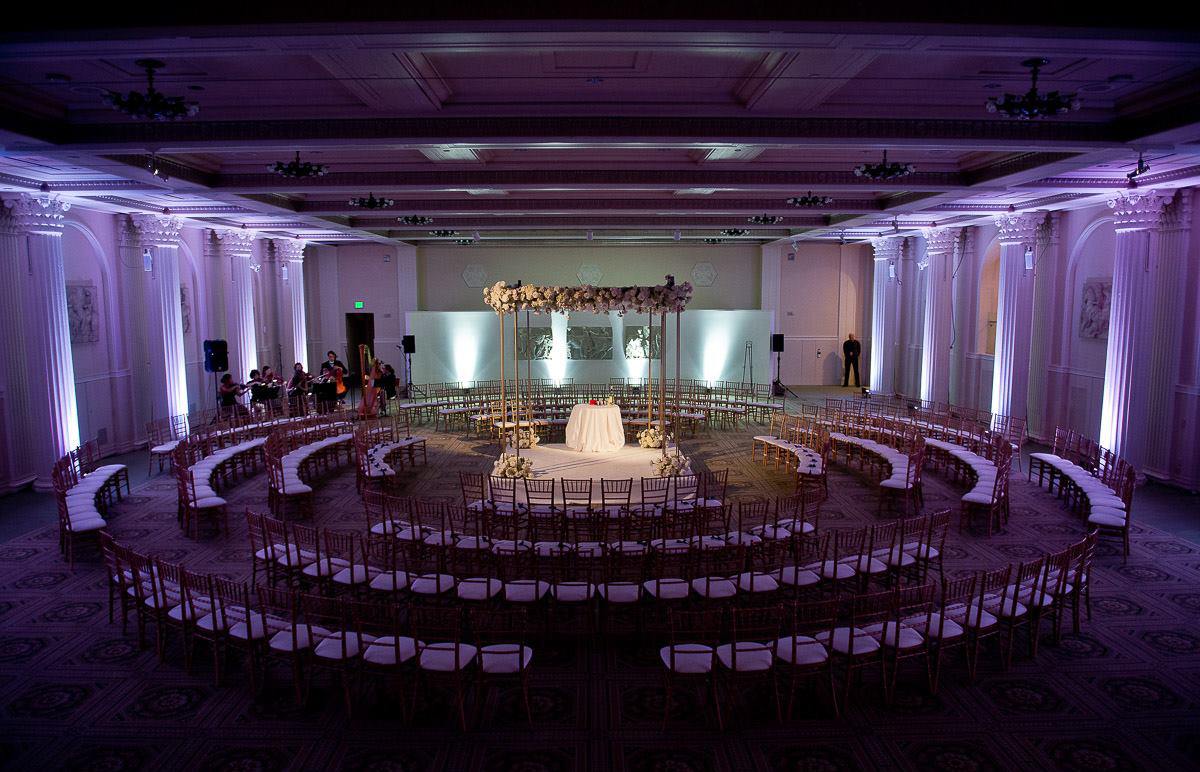 Color photograph of a dimly lit ballroom with chairs in circles around an illuminated stage for a ceremony in the middle. White Greek columns line both sides of the room.