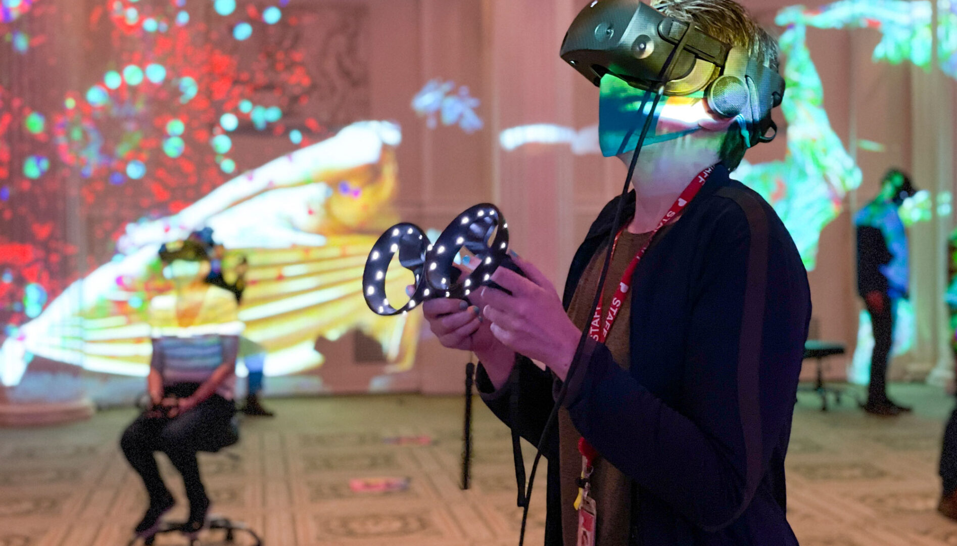 A person in the Sunken Ballroom wearing a VR headset and hand controls. Other people doing virtual reality and colored lights are in the background.