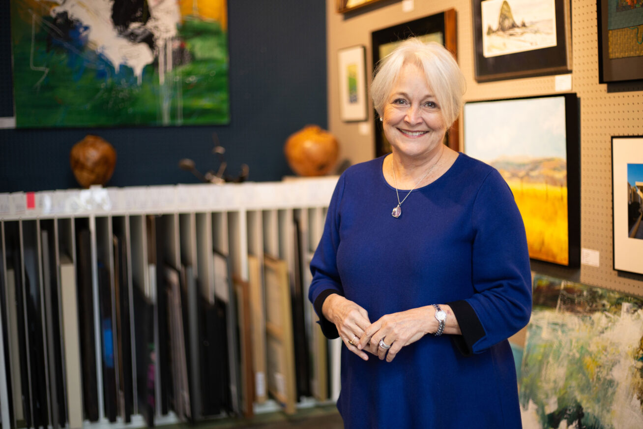 Woman with short white-blonde hair and a blue blouse, smiling with her hands folded in front of her. In the background are paintings hanging and in storage racks at the Rental Sales Gallery.
