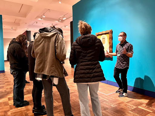 A man with a face mask on talking to a group of people in a gallery in front of a bright blue wall.
