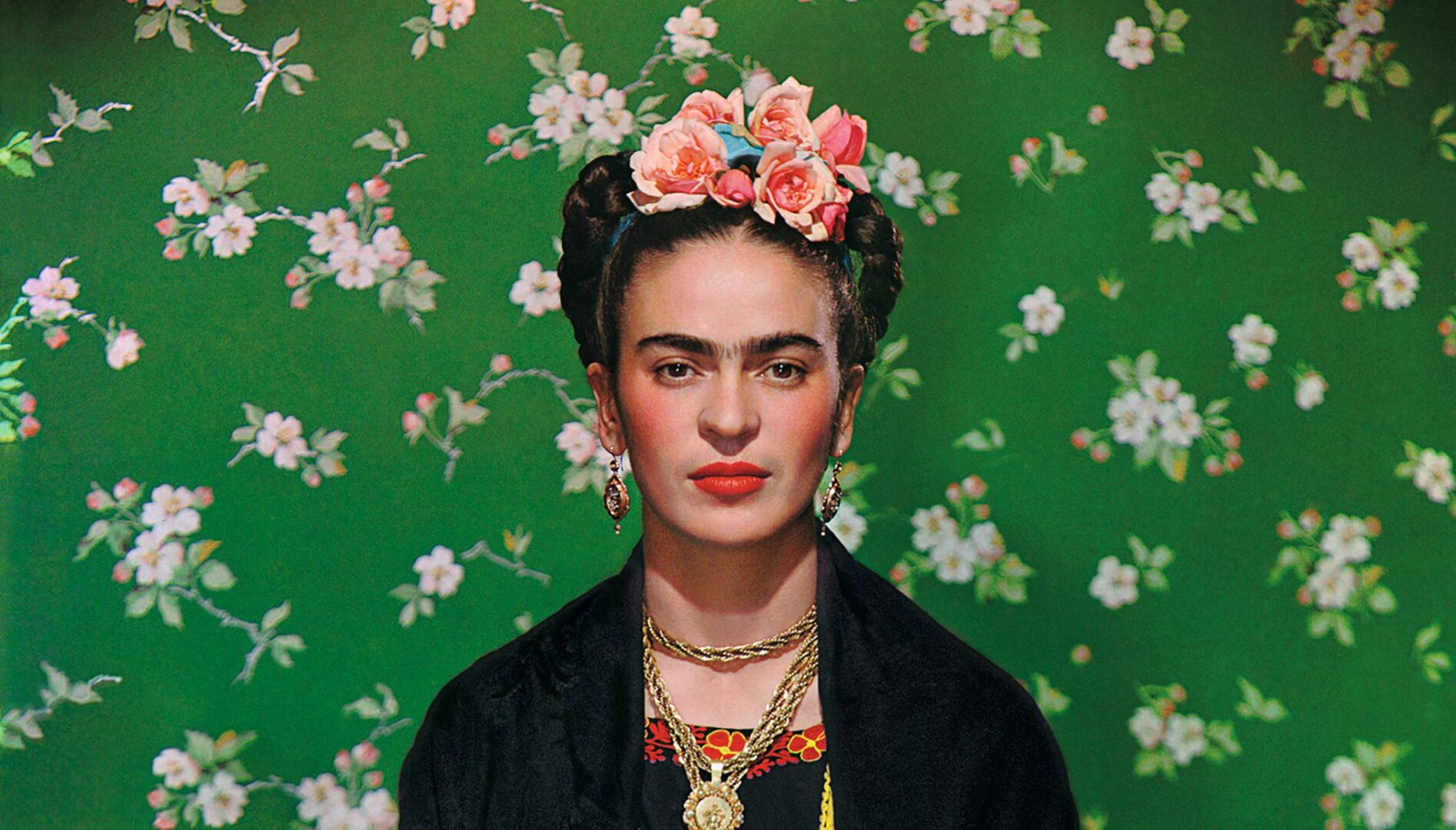 Frida Kahlo with a pink flower crown, heavy gold jewelry, and her hair up in braids, sitting against a bright green wallpaper with pink flowers on it.