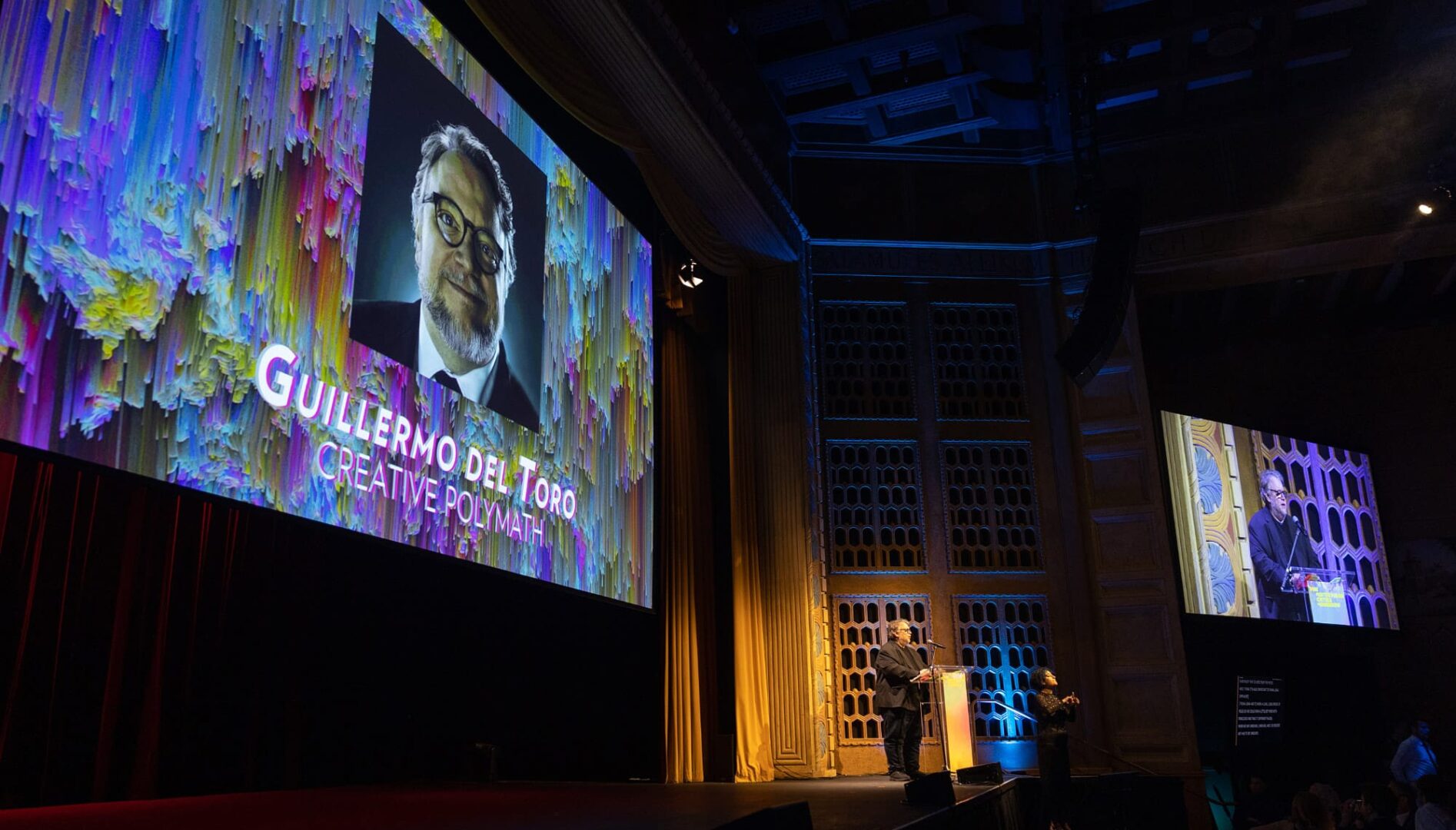 Guillermo del Toro speaking at podium with projections behind and to the side of him.