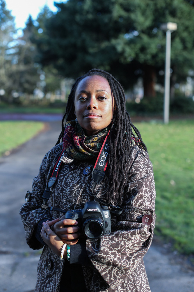 Photo of a Black woman with long hair in twists standing outside in a park with a camera around her neck and a brown and white paisley coat and scarf.