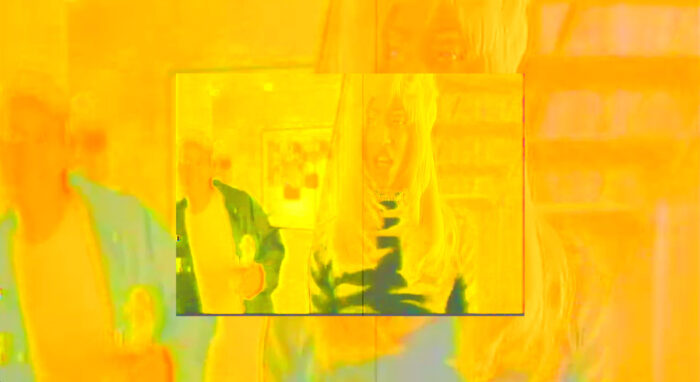 Video still of a rectangle within a larger rectangle, washed in yellow. A woman with a long blonde wig is in the foreground.