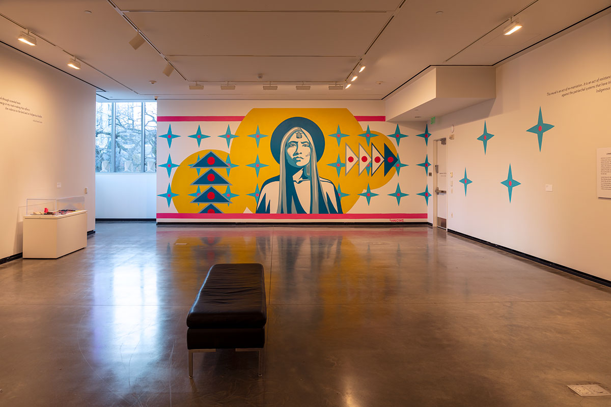 A gallery with a mural of a Native woman in the middle of yellow circles and blue stars and triangles with red circular centers